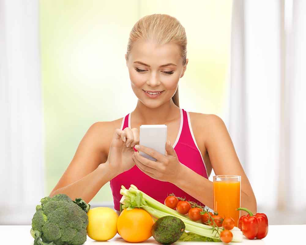 Fit woman sitting infront of fruits and veggies while counting macros using cellphone